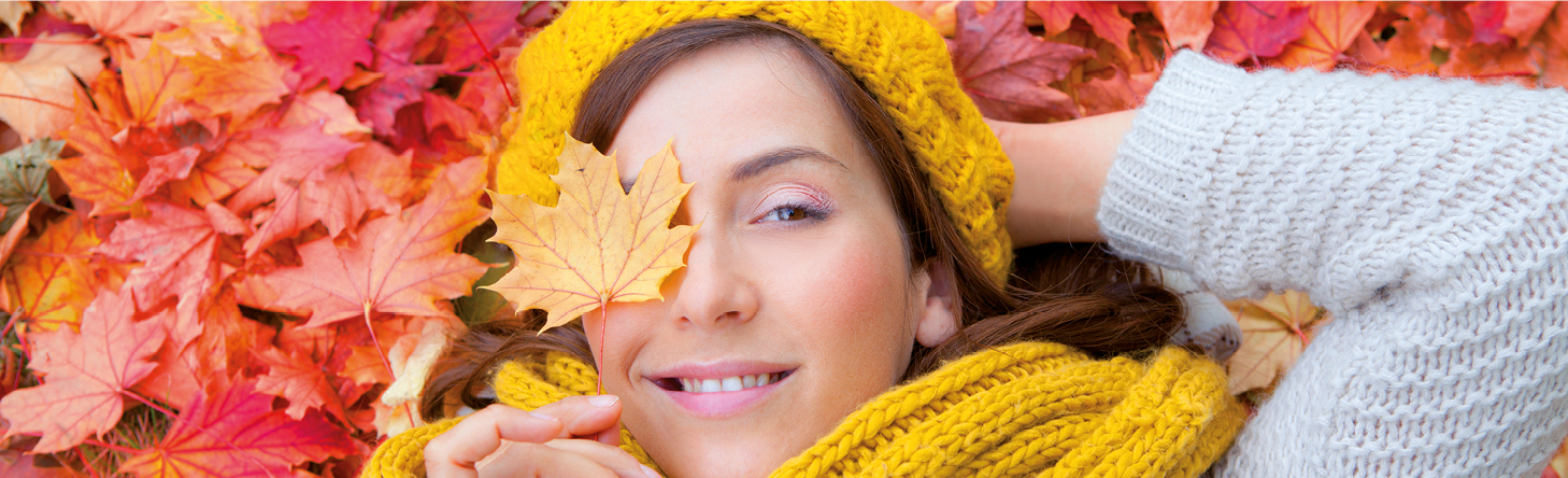 Auf einen Blick Frau mit gelber Mütze und gelbem Schal liegt im bunten Herbstlaub und hält sich ein gelbes Ahornblatt vor das Auge