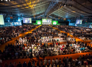 Eine Messehalle ist gefüllt mit vielen sitzenden Gästen, die ihren Blick nach Vorne zur Bühne richten, wo sich zwei Personen unterhalten