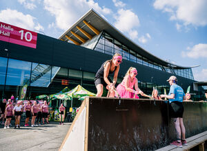 Zwei Frauen im Vordergrund überwinden ein Hindernis bei einem Hindernislauf, im Hintergrund stehen weitere Teilnehmerinnen