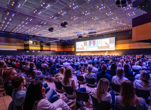 Blick von rechts auf eine Bühne und sitzende Gäste mit lila Beleuchtung
