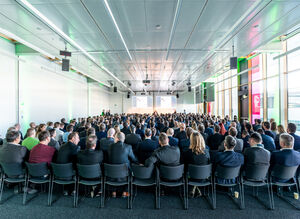 Blick von hinten auf einen voll belegten Tagungsraum und drei Sprechern im Hintergrund