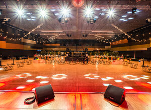 Blick von der Bühne auf die leere Location, zwei Servicekräfte bereiten die Tische vor