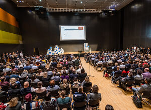 Vollbesetzter Raum mit sitzenden Gästen, die auf eine Bühne mit einer Rednerin blicken