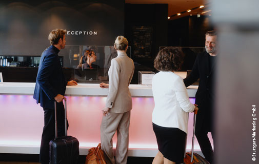 Vier Personen mit Rollkoffern stehen an der Rezeption eines modernen Hotels und sprechen mit der Rezeptionistin. Der Empfangstresen ist beleuchtet und die Umgebung wirkt hochwertig.