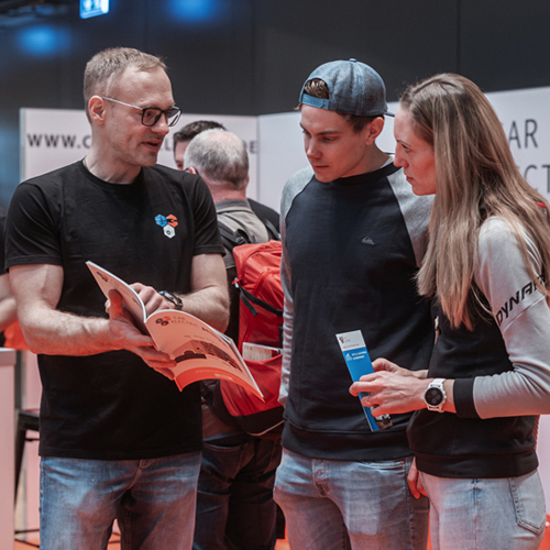 Ein Aussteller auf einer Messe erklärt einem jungen Paar Inhalte aus einer Broschüre.